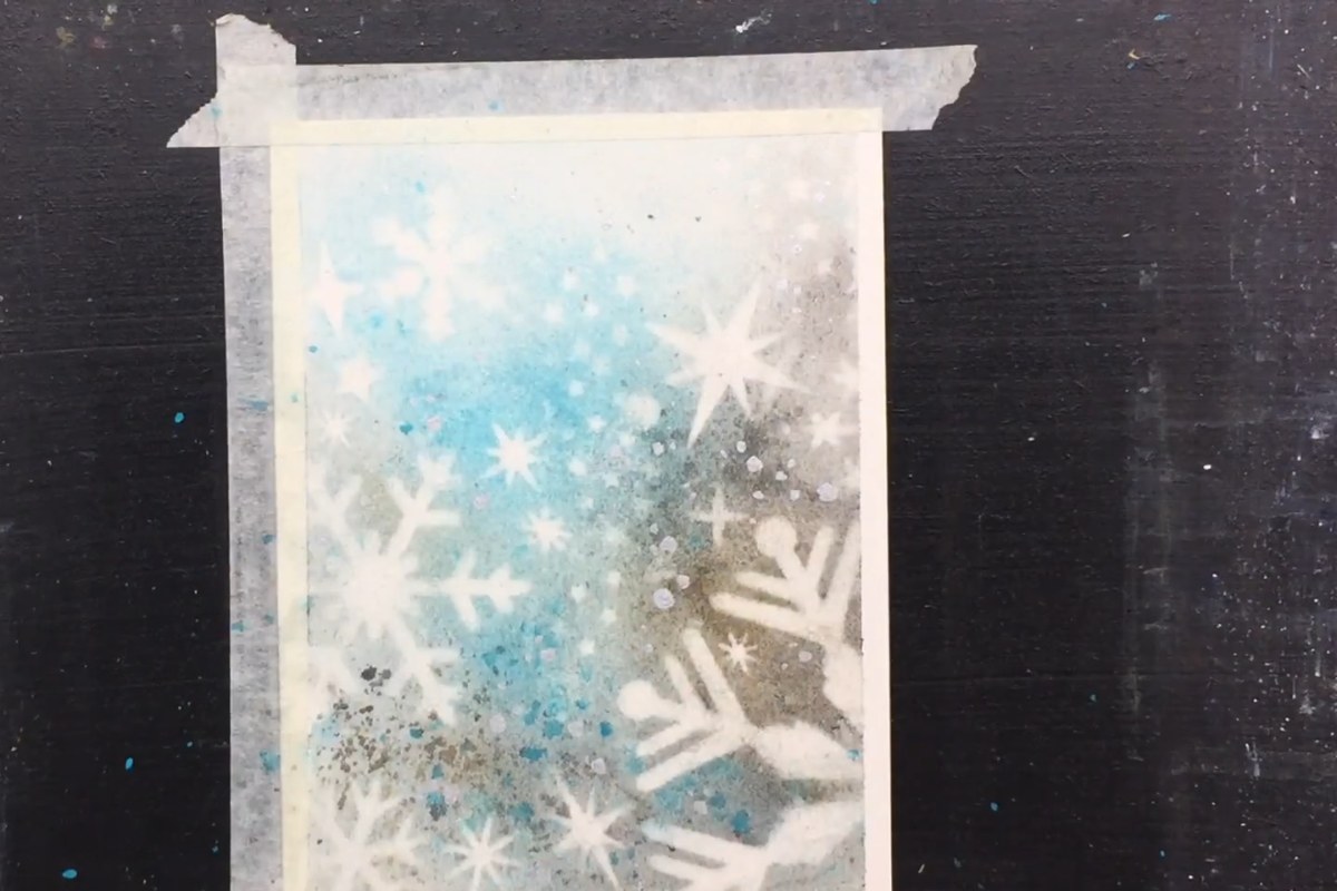 Peeling the tape off a blue and grey watercolor card with white stenciled snowflakes