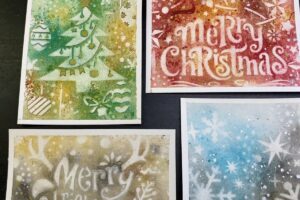 Four reverse stenciled watercolor Christmas cards: a green tree, a red Merry Christmas, a gold reindeer and a blue snowflake design